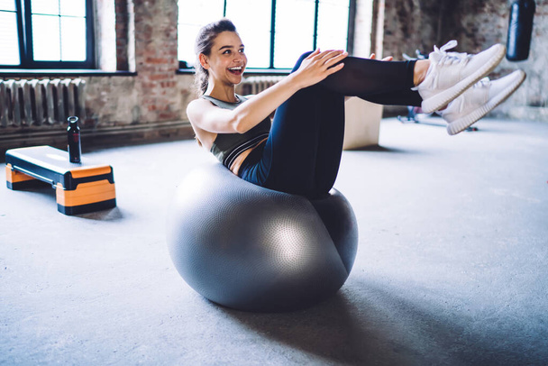 Full body of slim happy female in sportswear doing double crunches exercise on fit ball while having workout in spacious gym - Fotografie, Obrázek
