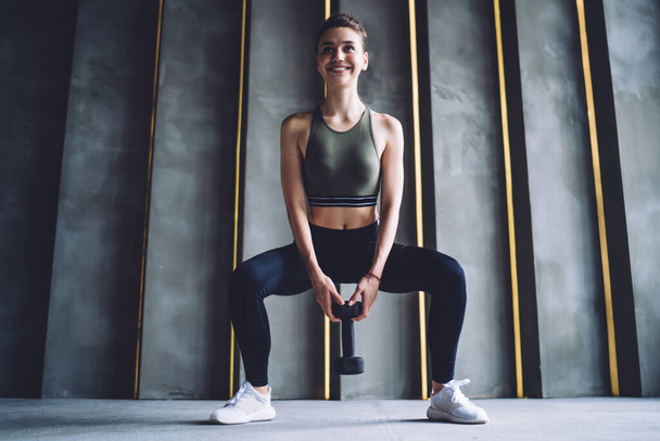 Full body of slim cheerful female in activewear standing on step platform with dumbbell and looking at camera during workout - Фото, зображення