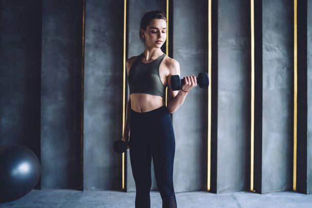 Fit female athlete in black sportswear performing exercises using black dumbbells while training biceps in modern fitness club against gray wall - Foto, Imagem