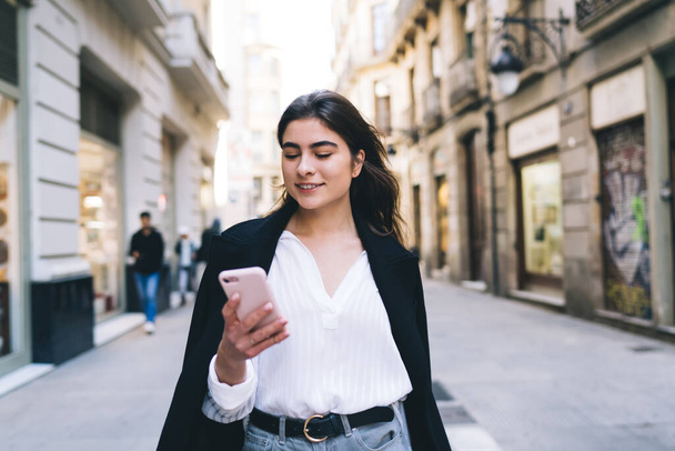 Positive stylish female in trendy outfit with toothy smile browsing smartphone while walking on pavement near old buildings after workday - 写真・画像