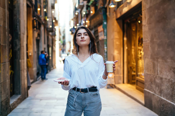 Stylish young female in casual clothes walking on old narrow street while drinking takeaway coffee and listening to music on smartphone - Fotó, kép