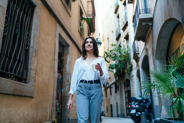 Low angle of cheerful female in casual clothes walking on street and drinking hot takeaway coffee while looking away and smiling - Zdjęcie, obraz