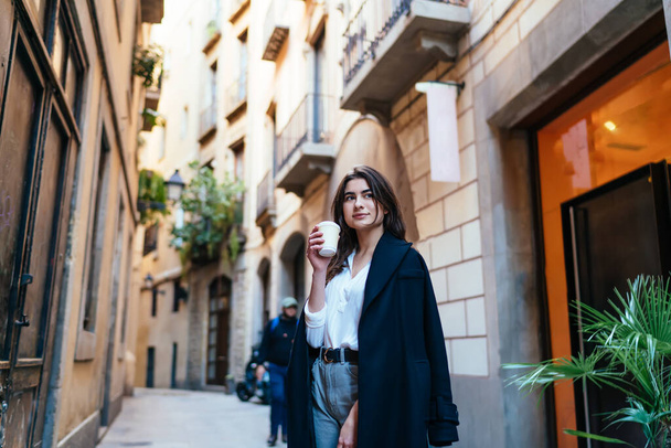 Pensive female tourist in trendy coat standing at street in historic center and pondering on old times, Caucasian hipster girl with disposable takeaway cup thinking during travel sightseeing - Photo, Image