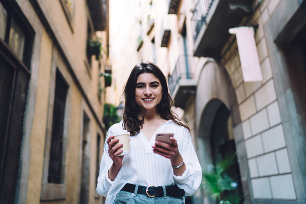 Half length portrait of pretty hipster girl with mobile smiling at camera during city sightseeing with coffee to go, carefree Caucasian blogger with modern smartphone gadget posing at touristic street - Foto, Imagen