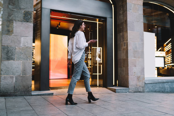 Side view of Caucasian female millennial dressed in casual clothing walking at urban street using cellphone app for listening music via electronic earphones, young hipster girl enjoying song records - 写真・画像