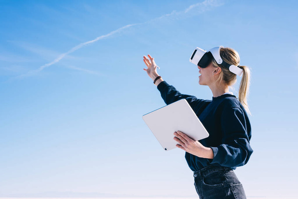 Side view of excited blond woman in casual clothes exploring virtual reality while using VR goggles and modern portable tablet in sunny day - Foto, afbeelding