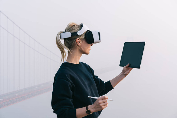 Side view of cheerful female freelancer holding digital tablet while wearing modern glasses virtial reality during work against misty bridge - Фото, зображення