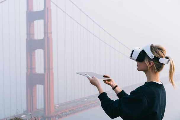 Side view of female with short blond hair in black sweater holding tablet and pencil while standing near bridge located in San Francisco and wearing VR headset - Fotó, kép