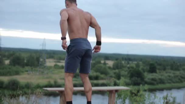 Un joven musculoso con un torso desnudo entrena junto al río y bombea los músculos de sus piernas mientras salta a un banco. Vista trasera. Primer plano - Imágenes, Vídeo