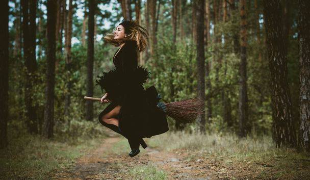 Young beautiful and mysterious Witch woman Flying on the Broom in woods. Levitation, a witch on a broom, free space. Halloween holiday, magic - Zdjęcie, obraz