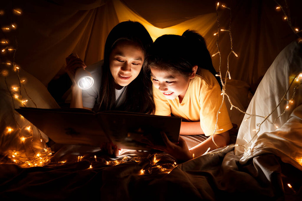 Family concept. Mother and child daughter reading book with flashlight together in children tent before bedtime. Happy mother read story book to her daughter in bed sheet ten - Photo, Image