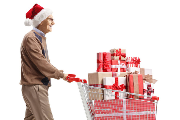 Elderly man with a santa claus hat pushing a shopping cart with christmas presents isolated on white background - Foto, Bild