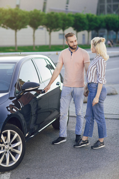 Mężczyzna daje kobiecie niespodziankę, kupując nowy samochód. Szczęśliwa młoda para w pobliżu nowego samochodu na zewnątrz. Kupowanie samochodu - Zdjęcie, obraz