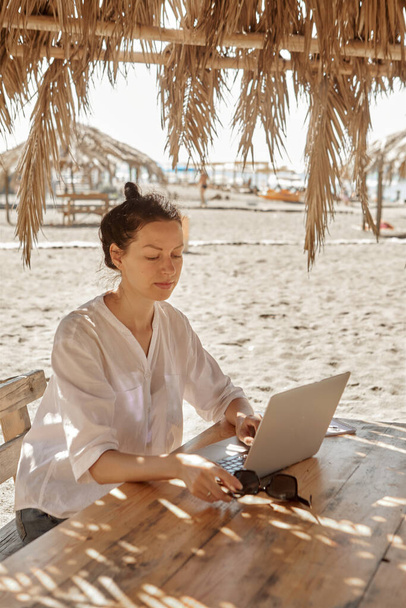 Kumsalda dizüstü bilgisayar kullanan genç bir kadın. Serbest çalışma kavramı - Fotoğraf, Görsel