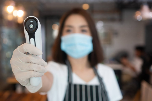 Asian waitress woman wearing face masks and holding an infrared forehead thermometer to check body temperature for virus symptoms of customers before entering the restaurant ( coffee shop ). - Fotó, kép