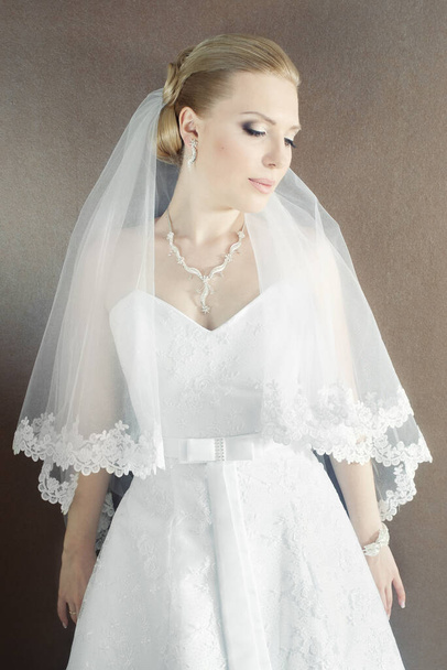 Όμορφη νύφη με λευκό φόρεμα και πέπλο κοιτάζοντας κάτω στον καφέ τοίχο - Φωτογραφία, εικόνα