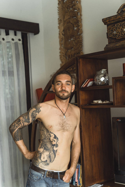 Young attractive man with a naked torso in tattoos at home. Portrait of a sexy man in a home atmosphere. - Valokuva, kuva