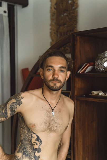 Young attractive man with a naked torso in tattoos at home. Portrait of a sexy man in a home atmosphere. - Foto, immagini