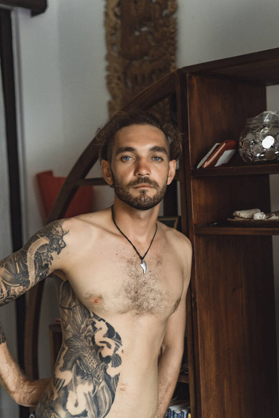 Young attractive man with a naked torso in tattoos at home. Portrait of a sexy man in a home atmosphere. - Valokuva, kuva