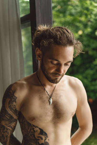 Young attractive man with a naked torso in tattoos at home. Portrait of a sexy man in a home atmosphere. - Фото, изображение