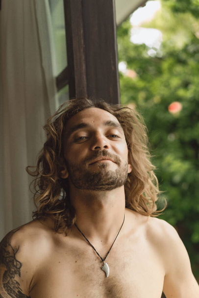 Young attractive man with long curly hair and naked torso at home. Portrait of a sexy man in a home atmosphere. - Foto, Imagem