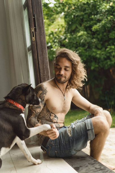 Young man with long curly hair and a naked torso at home. A man plays with a dog in a homely atmosphere in the yard. - Foto, immagini