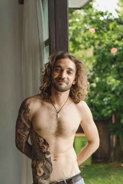 Young attractive man with a naked torso in tattoos at home. Portrait of a sexy man in a home atmosphere. - Фото, зображення