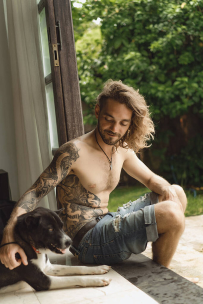 Young man with long curly hair and a naked torso at home. A man plays with a dog in a homely atmosphere in the yard. - Foto, afbeelding
