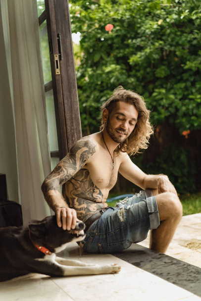 Young man with long curly hair and a naked torso at home. A man plays with a dog in a homely atmosphere in the yard. - Foto, imagen