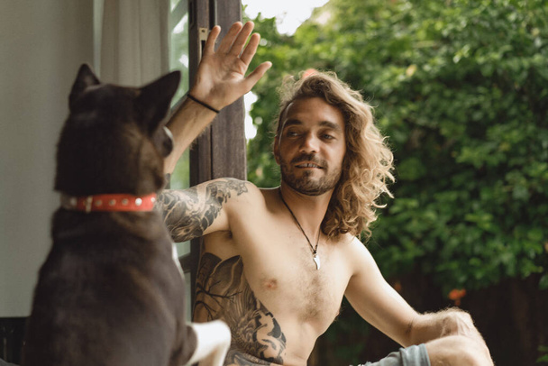 Young man with long curly hair and a naked torso at home. A man plays with a dog in a homely atmosphere in the yard. - Photo, image