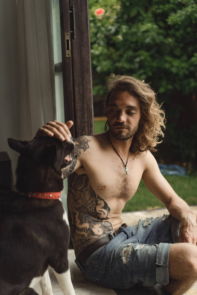 Young man with long curly hair and a naked torso at home. A man plays with a dog in a homely atmosphere in the yard. - Фото, изображение