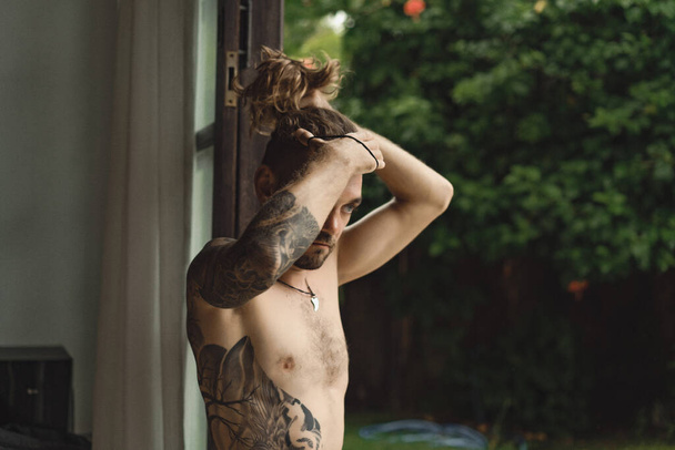 Young attractive man with long curly hair and naked torso at home. Portrait of a sexy man in a home atmosphere. - Фото, изображение