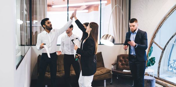 Group of positive multiethnic colleagues in formal clothes standing in modern office and giving high five to each other while celebrating triumph - Fotó, kép