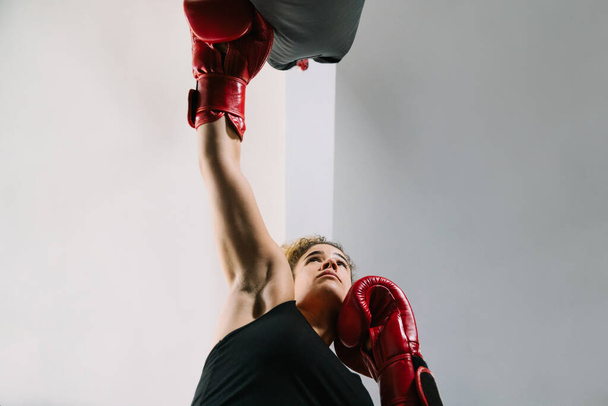 Kilátás lentről egy fehér lány piros bokszkesztyűt visel, amint egyenesen bokszzsákot üt egy edzőteremben. - Fotó, kép