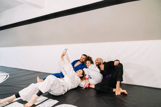 Judo gibi dört dövüş sanatları yoldaşının portresi kimono kullanıyor, spor salonunun duvarına yaslanıyor ve eğitimden sonra cep telefonuyla mutlu bir selfie çekiyor. - Fotoğraf, Görsel