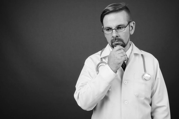 Studio πλάνο του ξανθό γενειοφόρος γιατρός άνθρωπος με μουσάκι σε γκρι φόντο σε μαύρο και άσπρο - Φωτογραφία, εικόνα