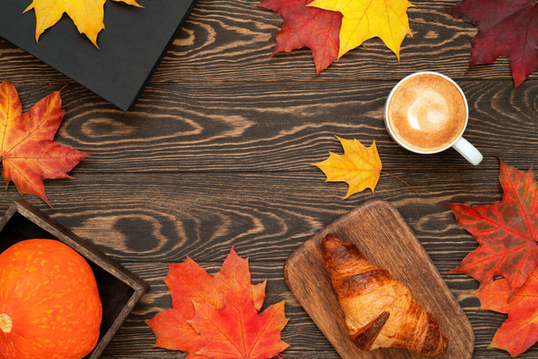 Autumn flat lay with cup of coffee, homemade croissant, book, pumpkin and colorful maple leaves on wooden table. Halloween background with copy space.  - Foto, Imagen