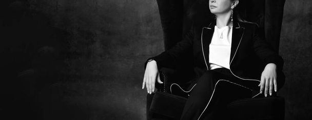 Black and white cropped photo of a woman in a black pantsuit sitting in a high chair in a comfortable position with one leg over the other. Cropped shot with dark gray background. - Foto, Imagem