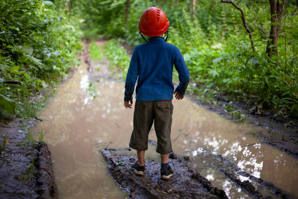 Child's Boy stands by a large puddle in the park. A child in a construction helmet. Lost son.  - Φωτογραφία, εικόνα