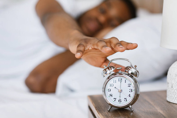 Αφρικανός ξυπνάει και κλείνει το ξυπνητήρι ξαπλωμένος στο κρεβάτι. - Φωτογραφία, εικόνα