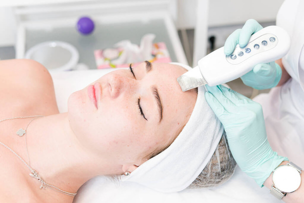 De vrouwelijke cosmetoloog maakt een procedure van ultrasone reiniging gezicht, close-up. Jonge vrouw in een schoonheidssalon. Begrip kosmetologie en professionele huidverzorging - Foto, afbeelding