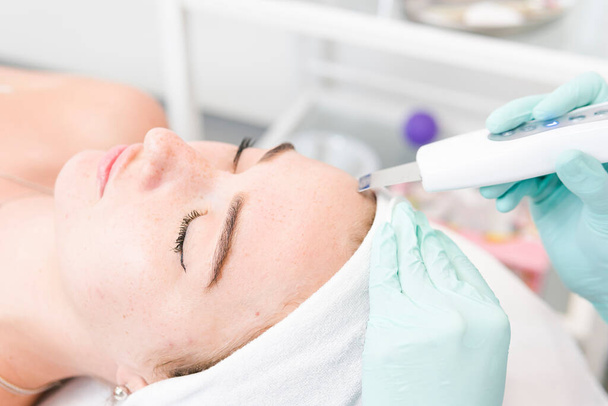 O cosmetologist feminino faz um procedimento da cara de limpeza ultrasônica, close-up. Uma jovem num salão de beleza. Conceito de cosmetologia e cuidados com a pele profissional - Foto, Imagem