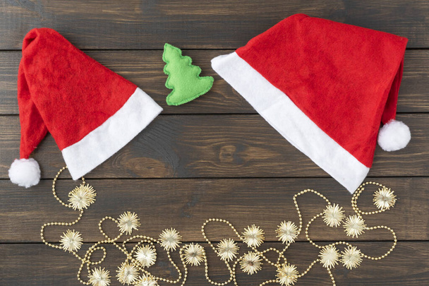 Auf dunklem Holzgrund liegen zwei Weihnachtsmützen und goldglitzernder Neujahrsschmuck. Kopieren Sie Platz für Text. Weihnachts- und Neujahrskonzept. - Foto, Bild
