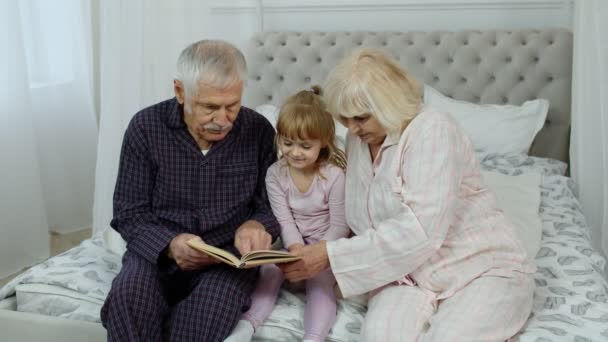 Cute girl with senior retired grandmother and grandfather sitting on bed reading book in bedroom - Footage, Video