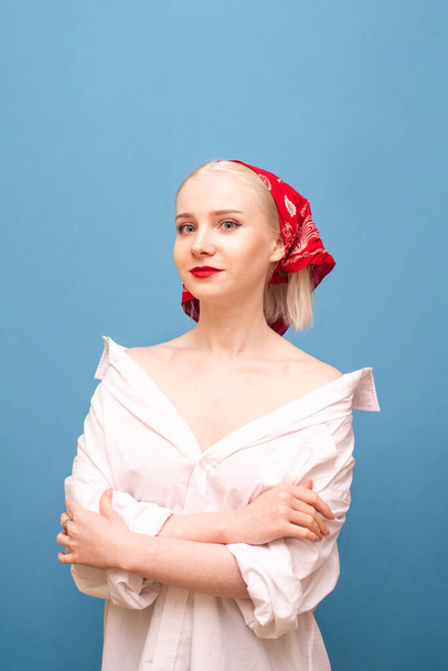 Cute blonde in headscarf and white men's shirt stands against a blue background, looks into the camera with a slight smile on her face. Vertical photo. Portrait of peasant woman. - Φωτογραφία, εικόνα
