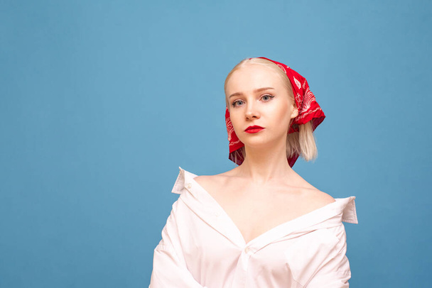 Portrait of a serious attractive blonde in a dress shirt and with a red scarf on her head, looks into the camera with a serious face. Pretty woman in headscarf and oversize shirt posing at camera. - Photo, Image