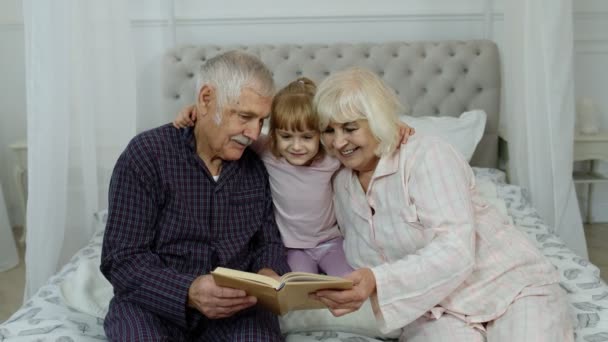 Cute girl with senior retired grandmother and grandfather sitting on bed reading book in bedroom - Footage, Video