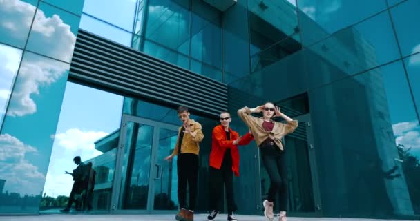Trzech młodych nastolatków w stylowych, jasnych koszulach i okularach przeciwsłonecznych pozujących przed kamerą w ruchu, poruszających się z wdziękiem, pokazujących odzież modną, dwóch facetów z dziewczyną tańczących na ulicy - Materiał filmowy, wideo