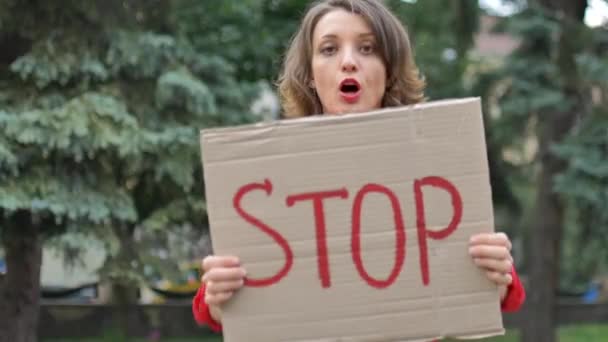 Kırmızı tişörtlü genç protestocu kadın protesto pankartı tutuyor. Üzerinde slogan olan tabela levhası. Ağaçların arkasındaki halk gösterisi için durun.. - Video, Çekim