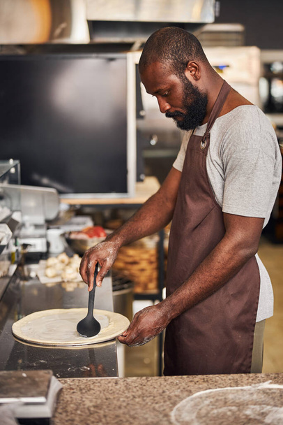 Young bearded man making pizza in cafe kitchen - Foto, afbeelding
